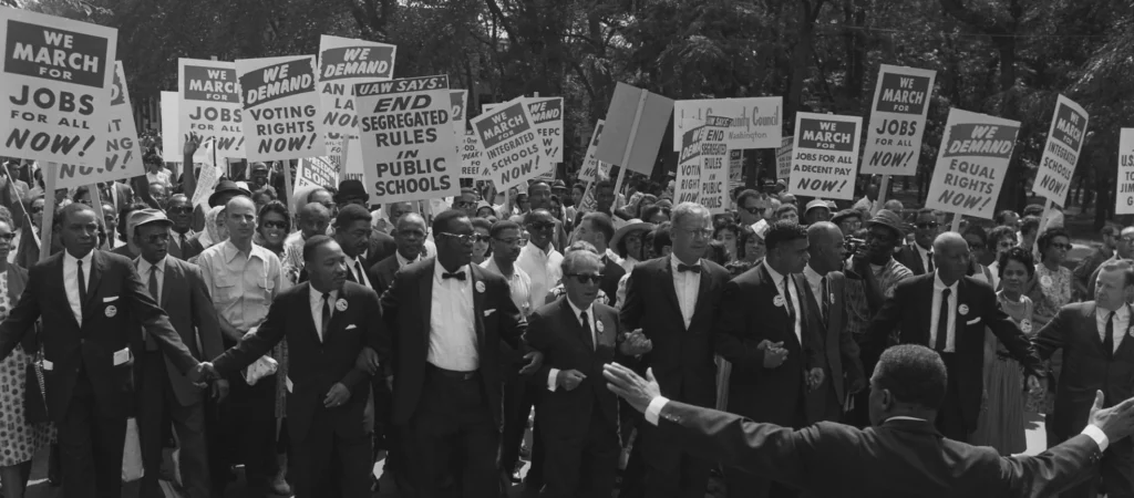 Historical black and white photograph of leaders at the March on Washington, D.C. on August 28, 1963, with protesters holding signs demanding jobs and civil rights